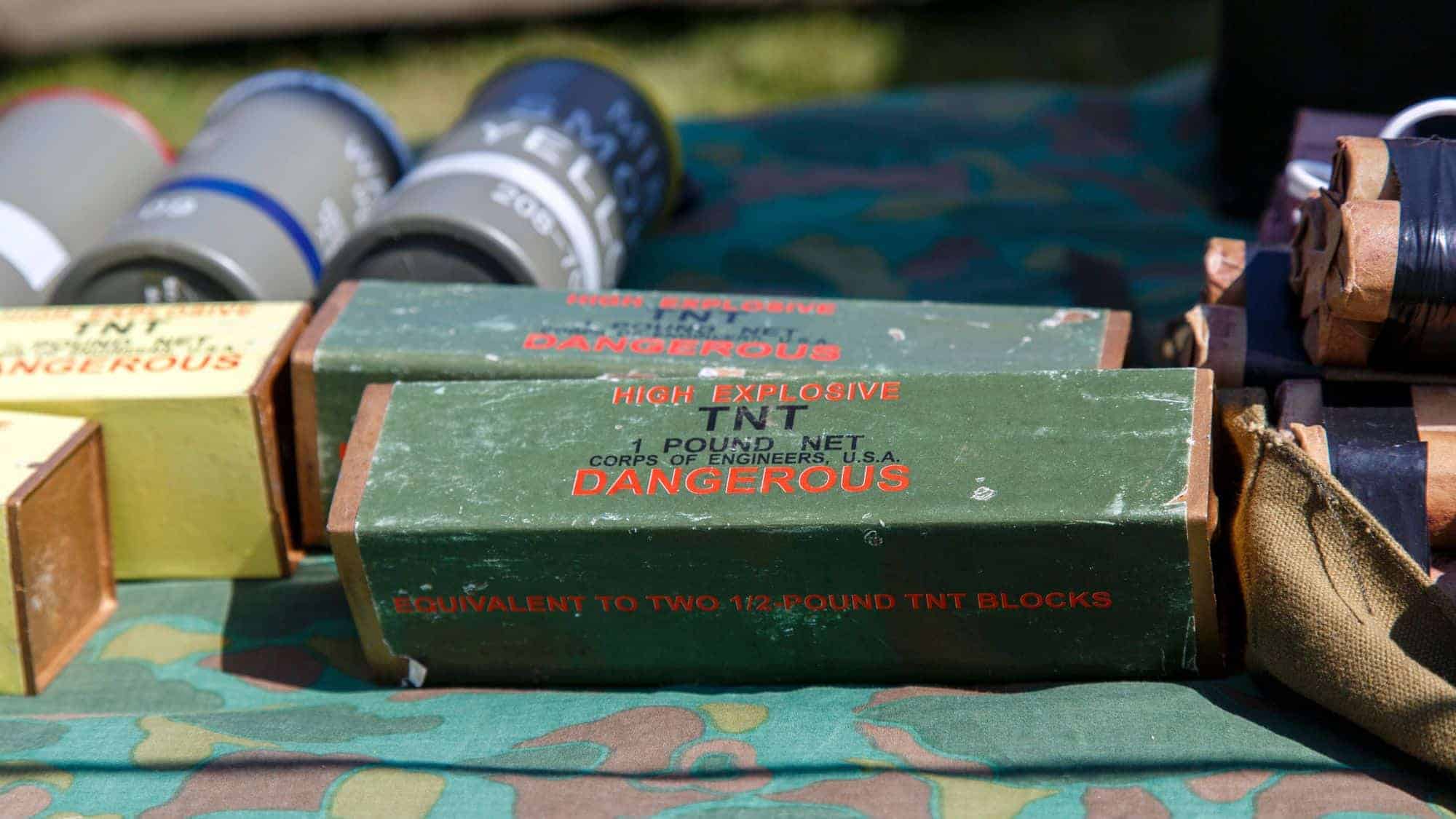 A collection of TNT explosives in green boxes labeled "High Explosive TNT 1 Pound Net" with warnings in red, displayed on a table with other military supplies.