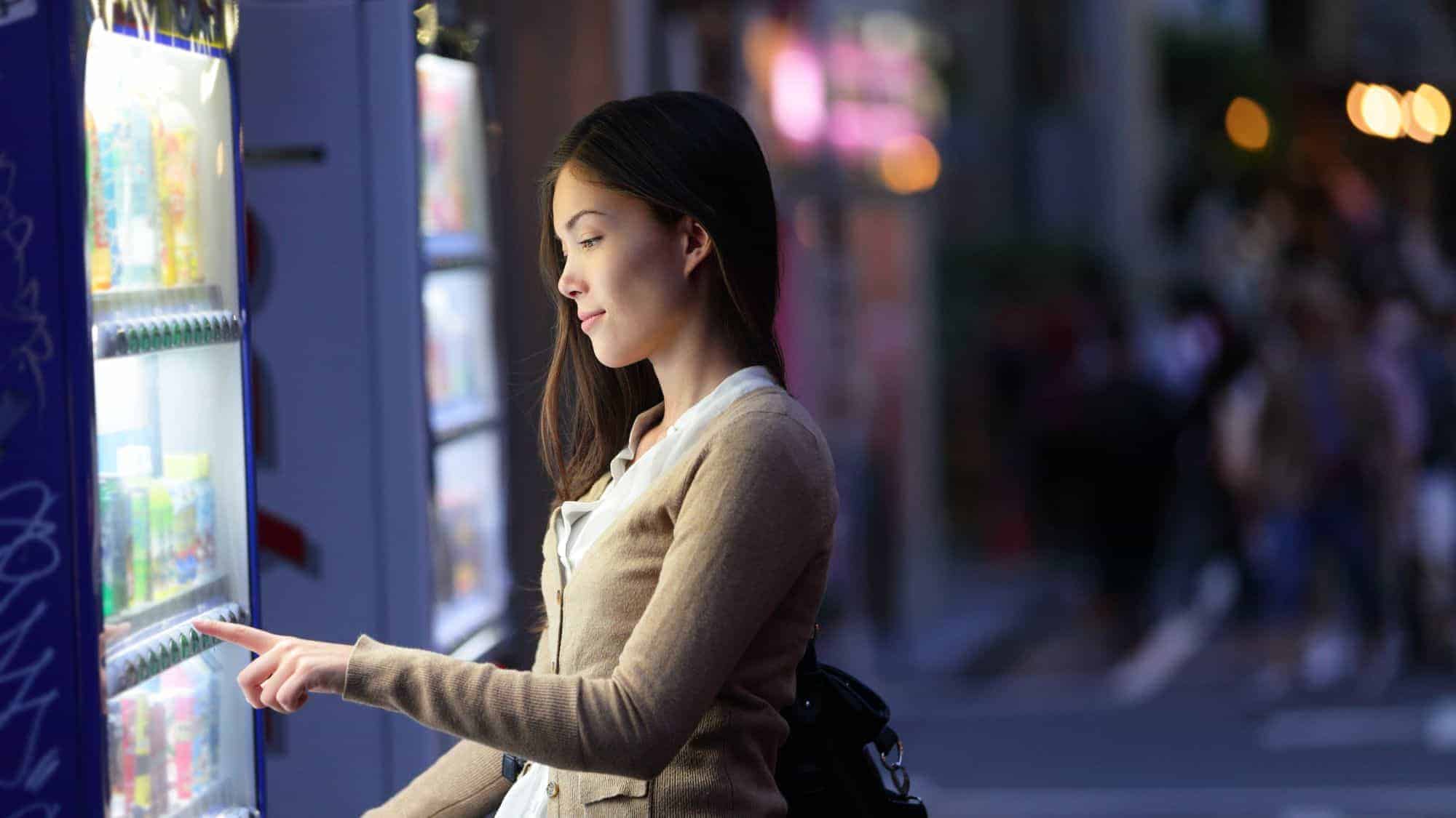 A woman selecting a product from a vending machine in a nighttime city setting, with bright lights illuminating the machine.