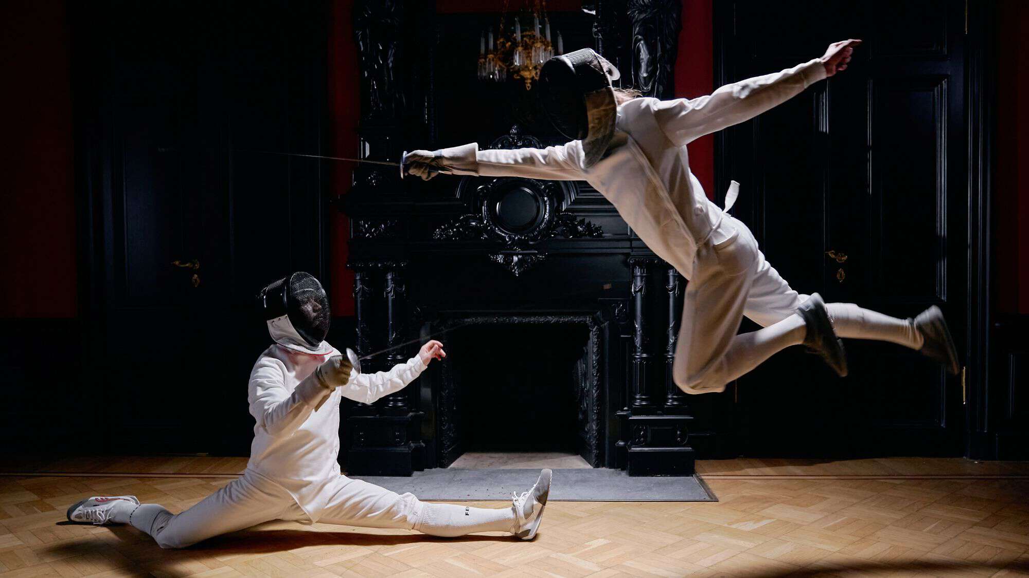 Two fencers mid-action, one leaping through the air while the other is in a split stance, both dressed in white fencing uniforms and masks in an elegant, dark room.