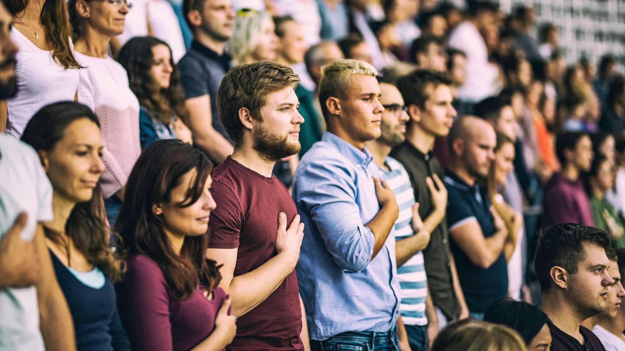 A crowd of people standing in a stadium, many with hands over their hearts, possibly during a national anthem or pledge of allegiance.