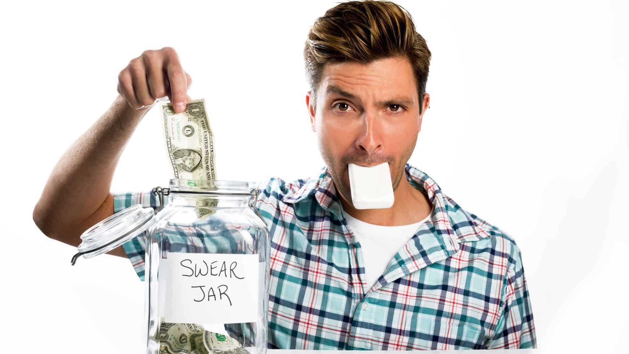 A man holding a dollar bill above a jar labeled "Swear Jar," while he has a bar of soap in his mouth.