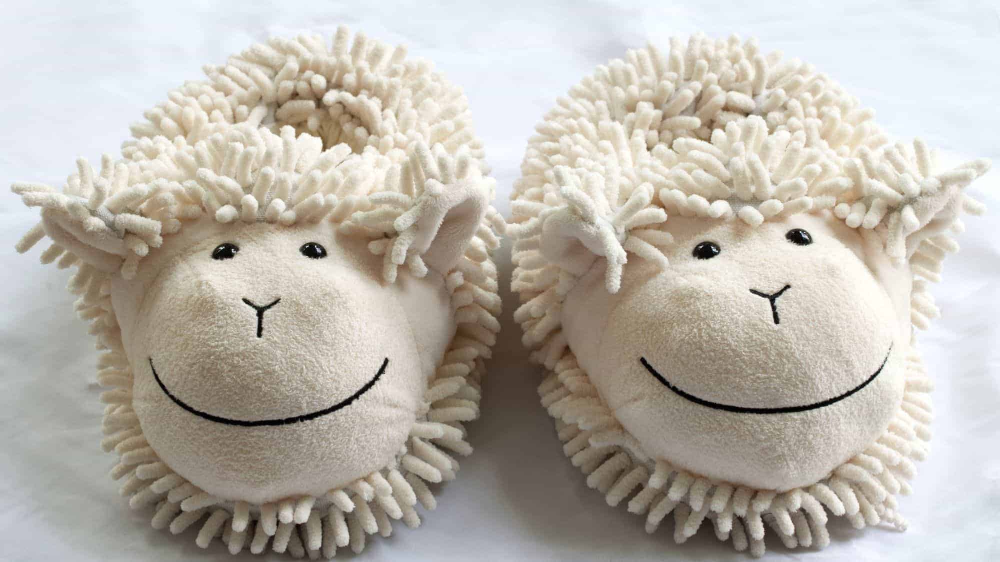 A pair of cute and fluffy sheep-themed slippers with smiling faces on a white surface.