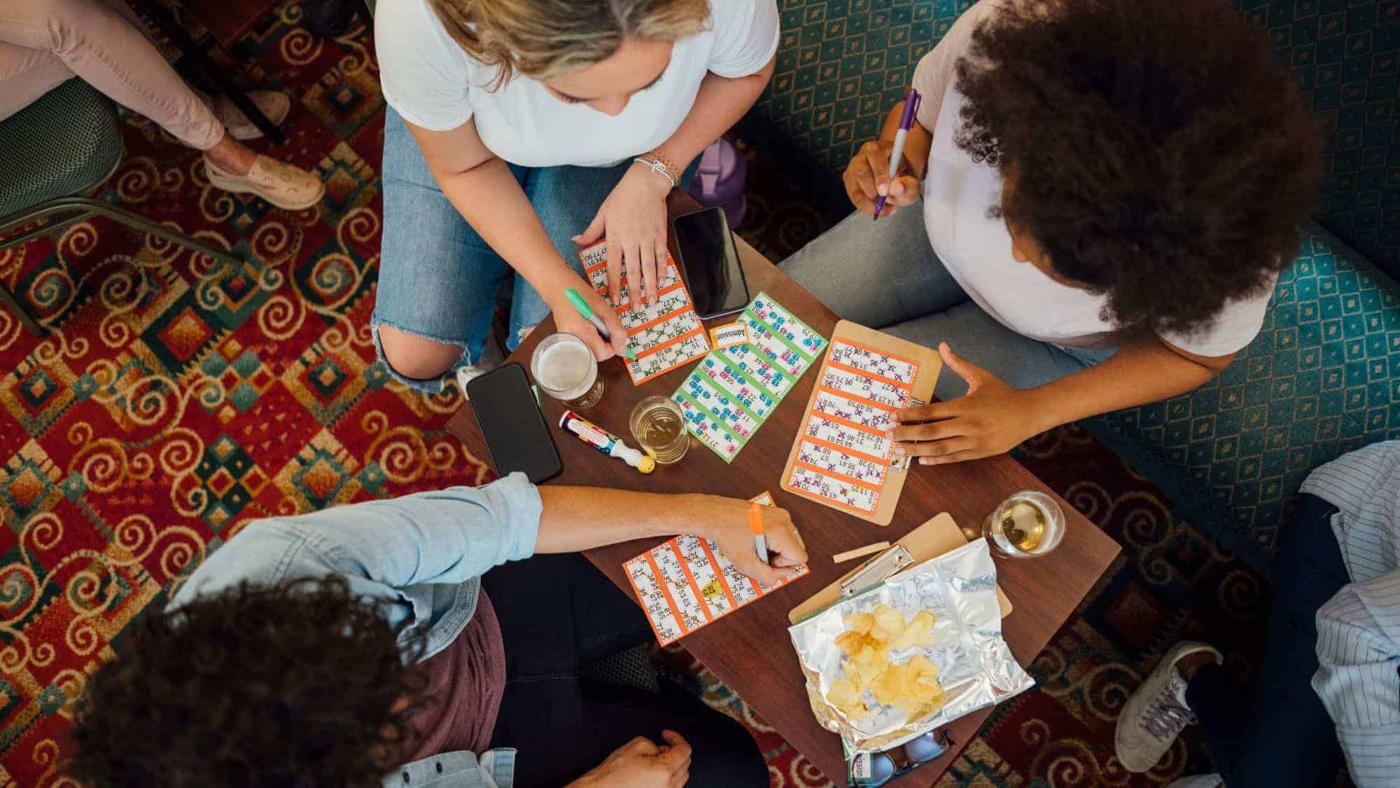 Overhead view of people sitting around a table playing a bingo game, marking their cards with markers, with drinks and snacks in the background.