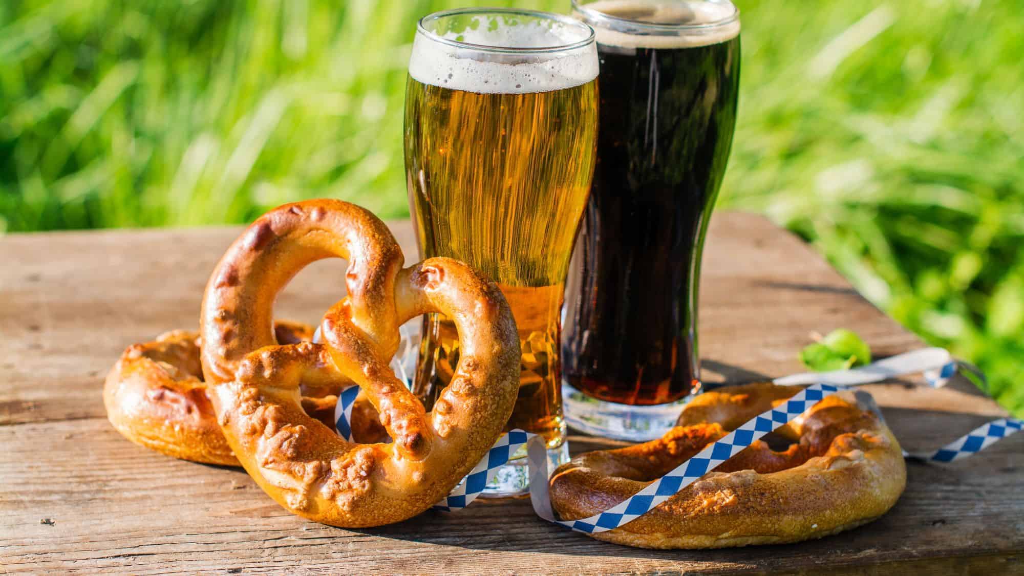 Two glasses of beer, one light and one dark, paired with large soft pretzels on a rustic wooden surface outdoors.