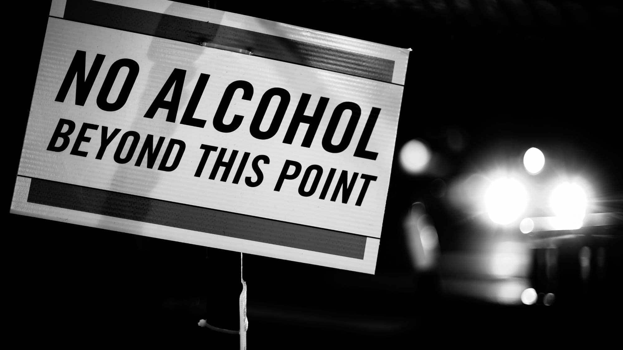 A black and white sign reading "No Alcohol Beyond This Point" with blurry headlights in the background at night.