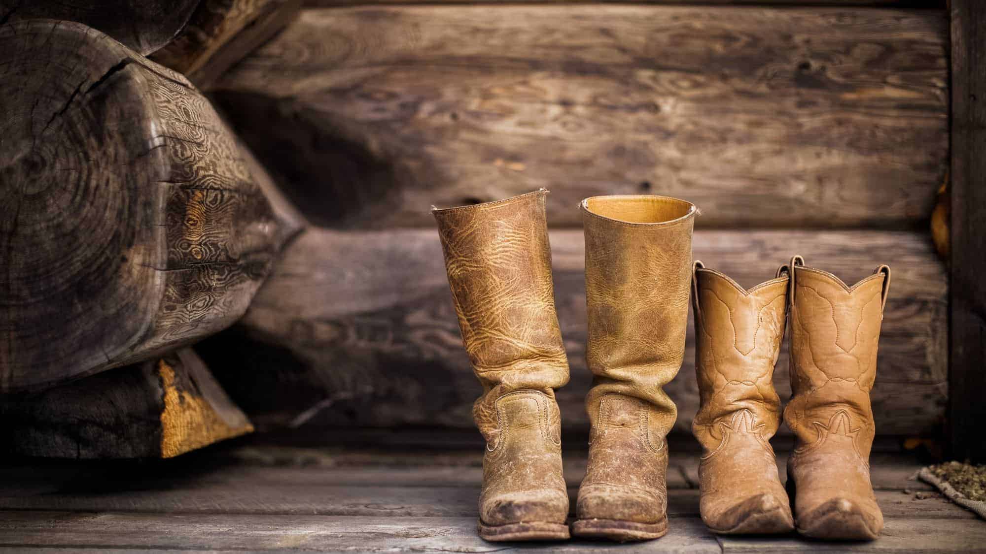 A set of old, worn cowboy boots, including one pair of adult-sized and one pair of child-sized, leaning against a wooden log wall.