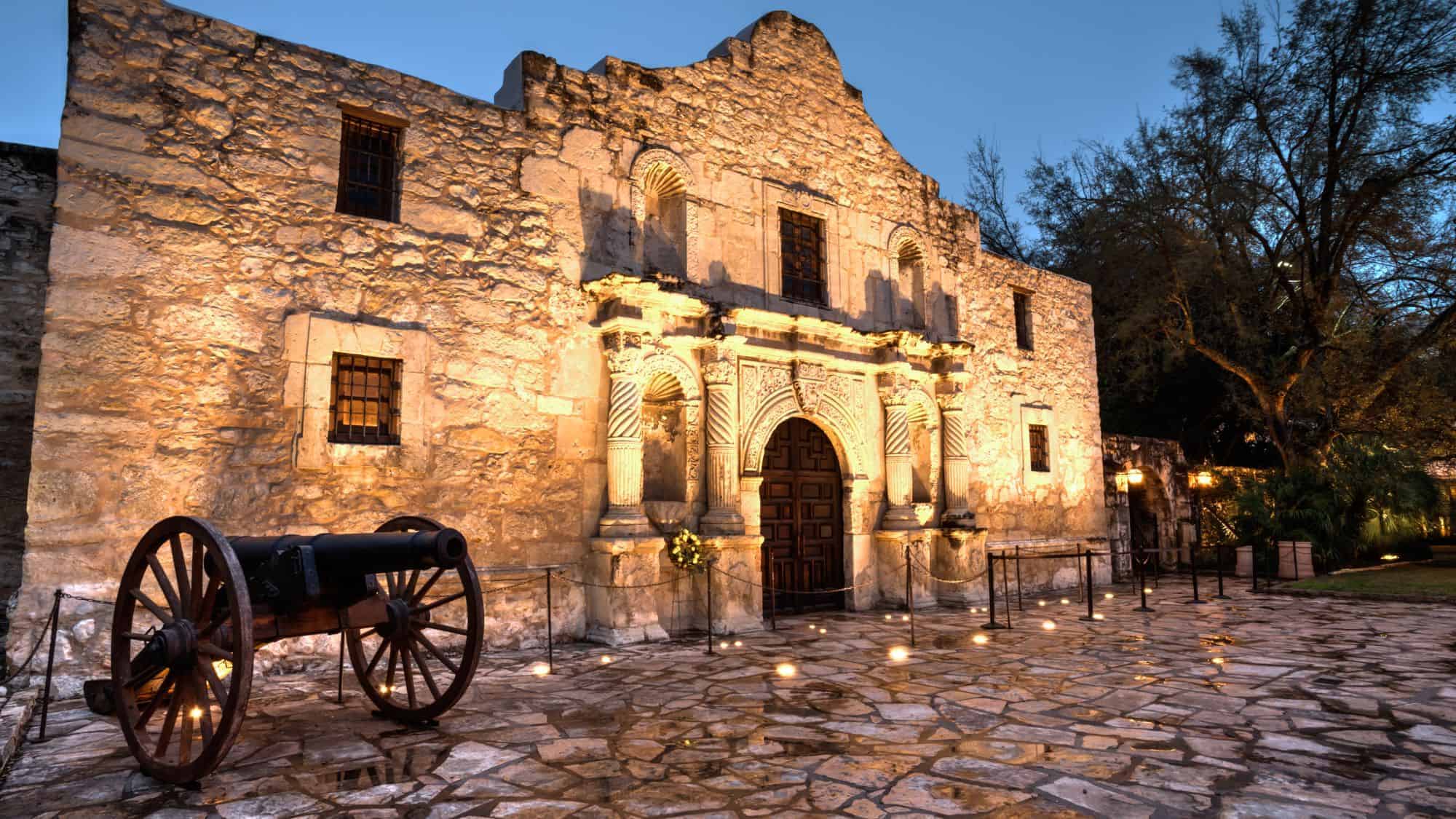A historic stone building, lit by warm lights, with a cannon in front of the entrance, likely depicting the Alamo in San Antonio.