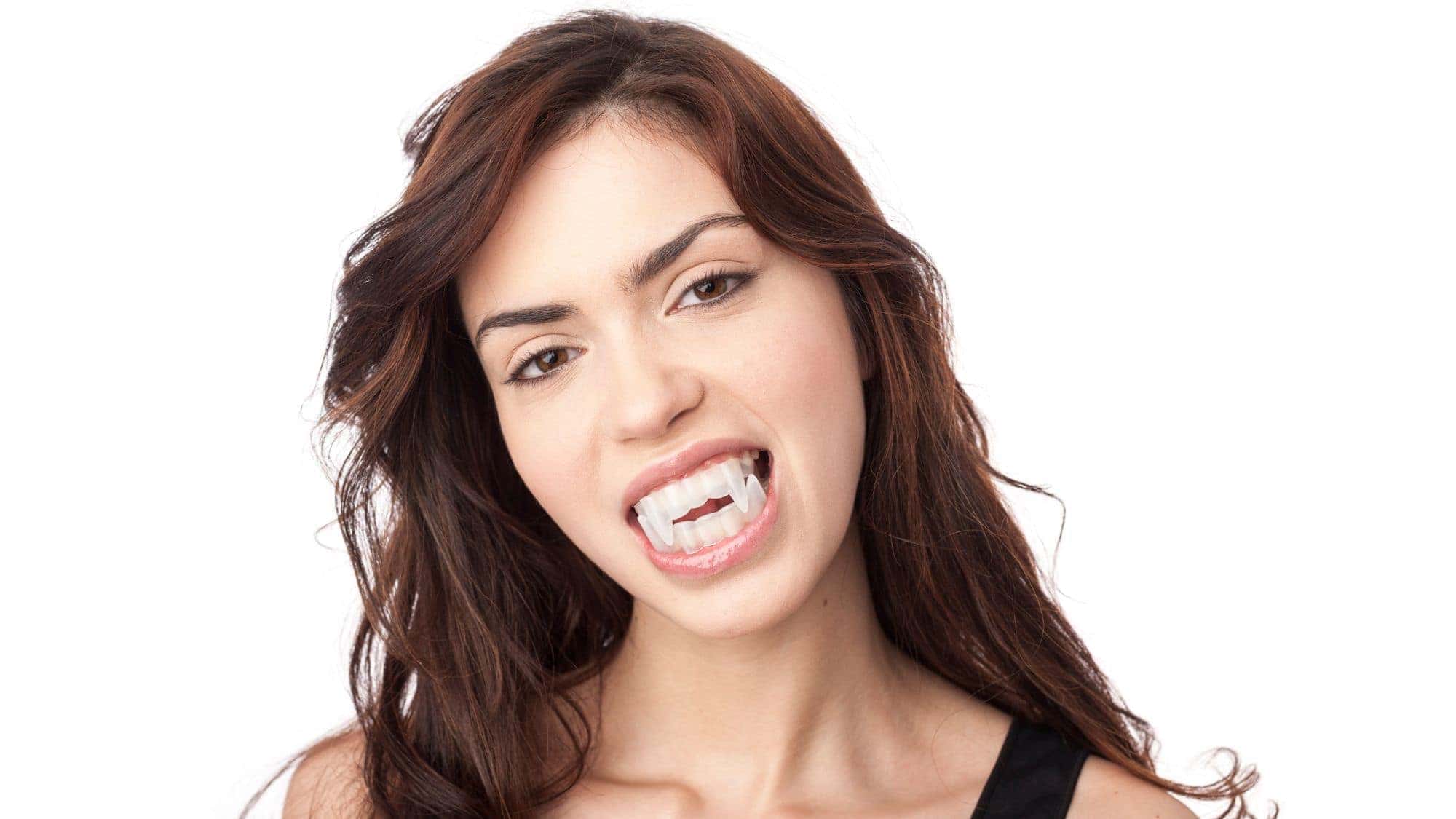 A woman with long hair wearing vampire fangs, looking playfully at the camera.