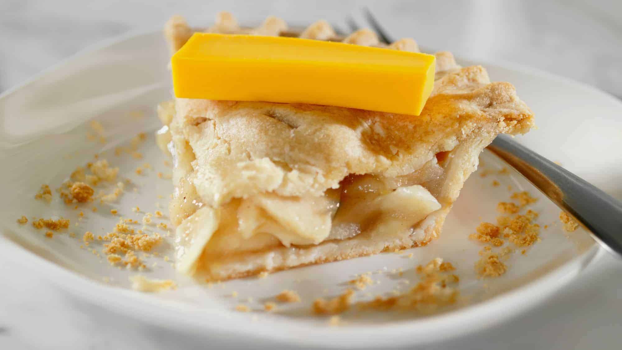 A slice of apple pie topped with a slice of cheddar cheese, resting on a white plate with some crumbs scattered around.