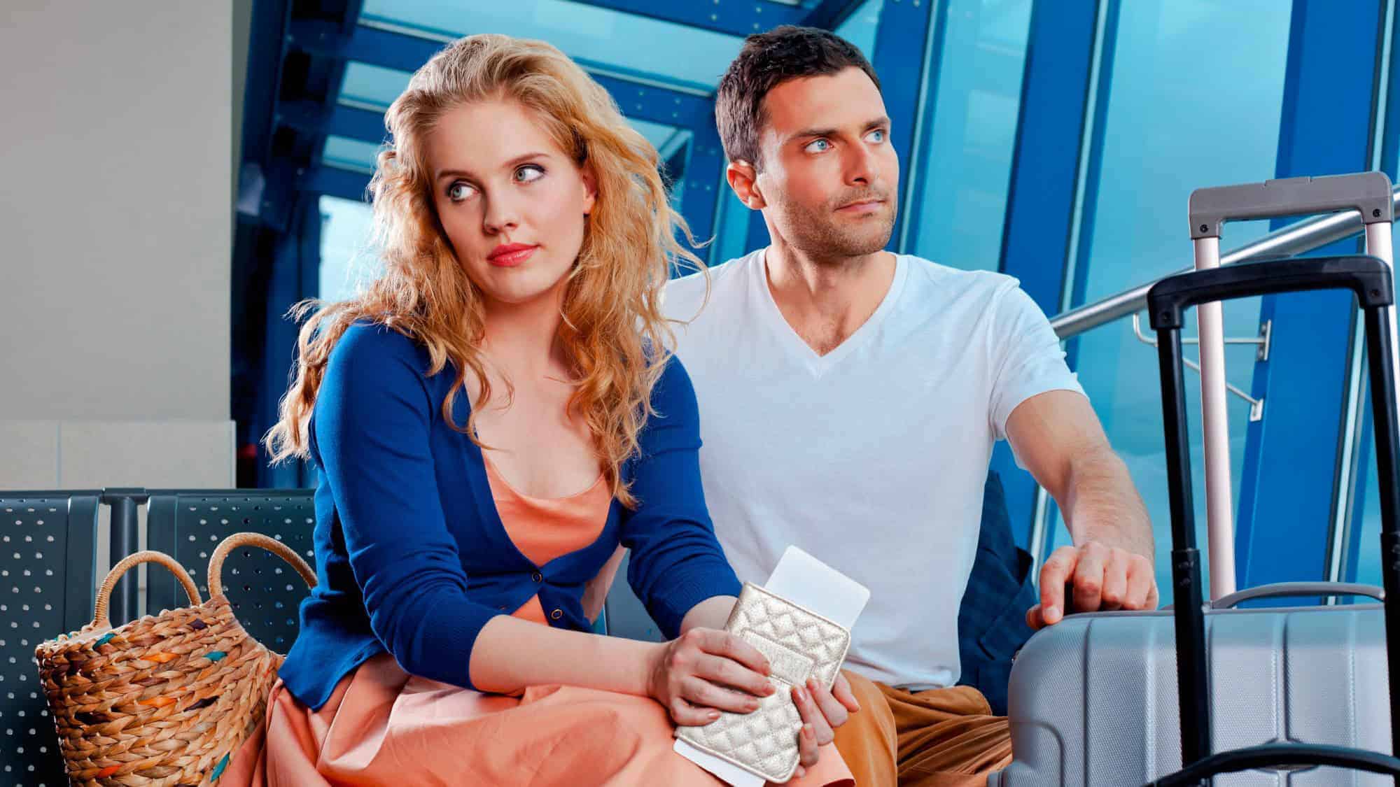 A frustrated couple sits in an airport, holding travel documents, as they wait for a delayed flight, with their luggage beside them.