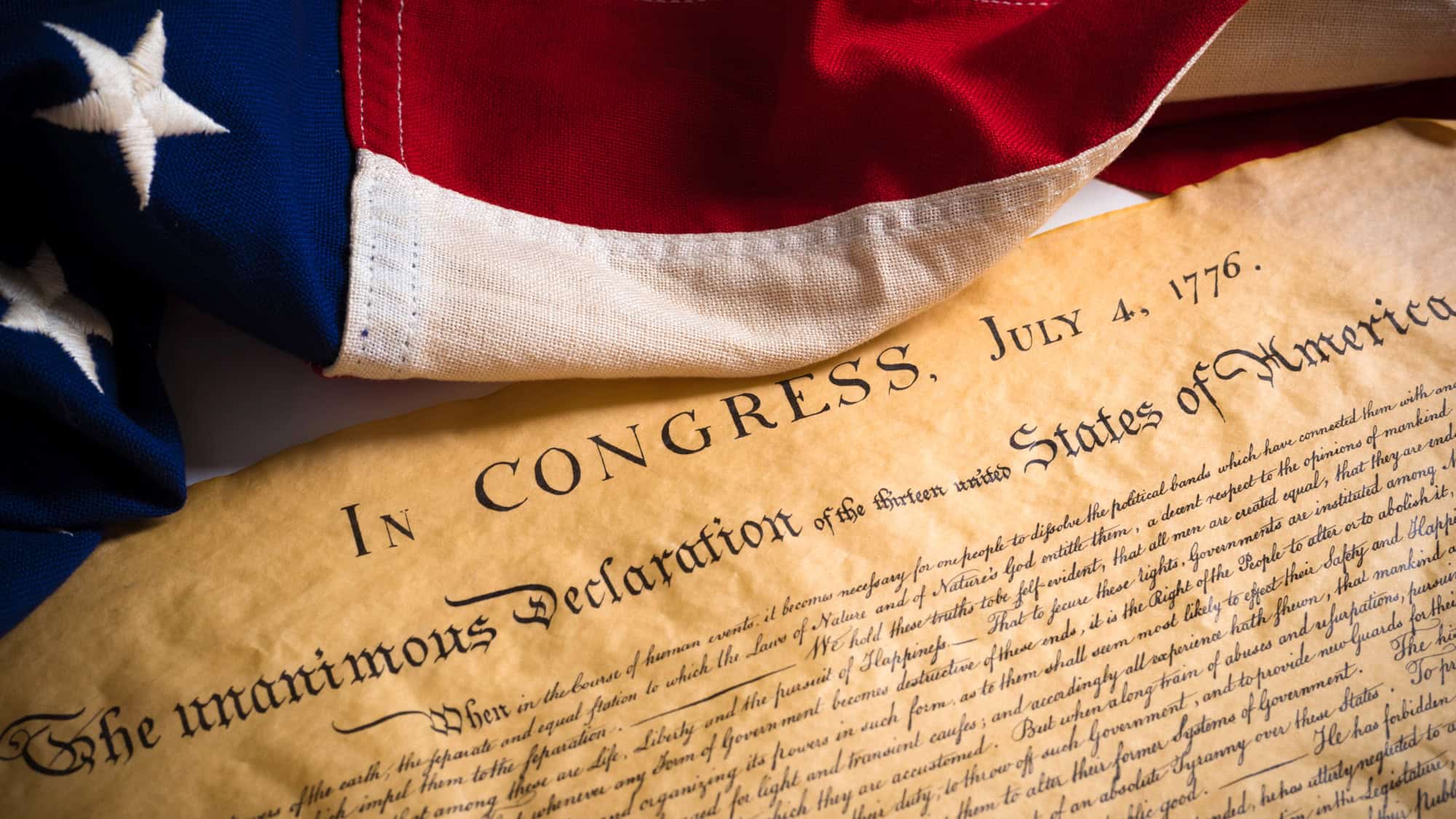 A close-up of the Declaration of Independence with a folded American flag draped above it, emphasizing the historical significance of July 4, 1776.