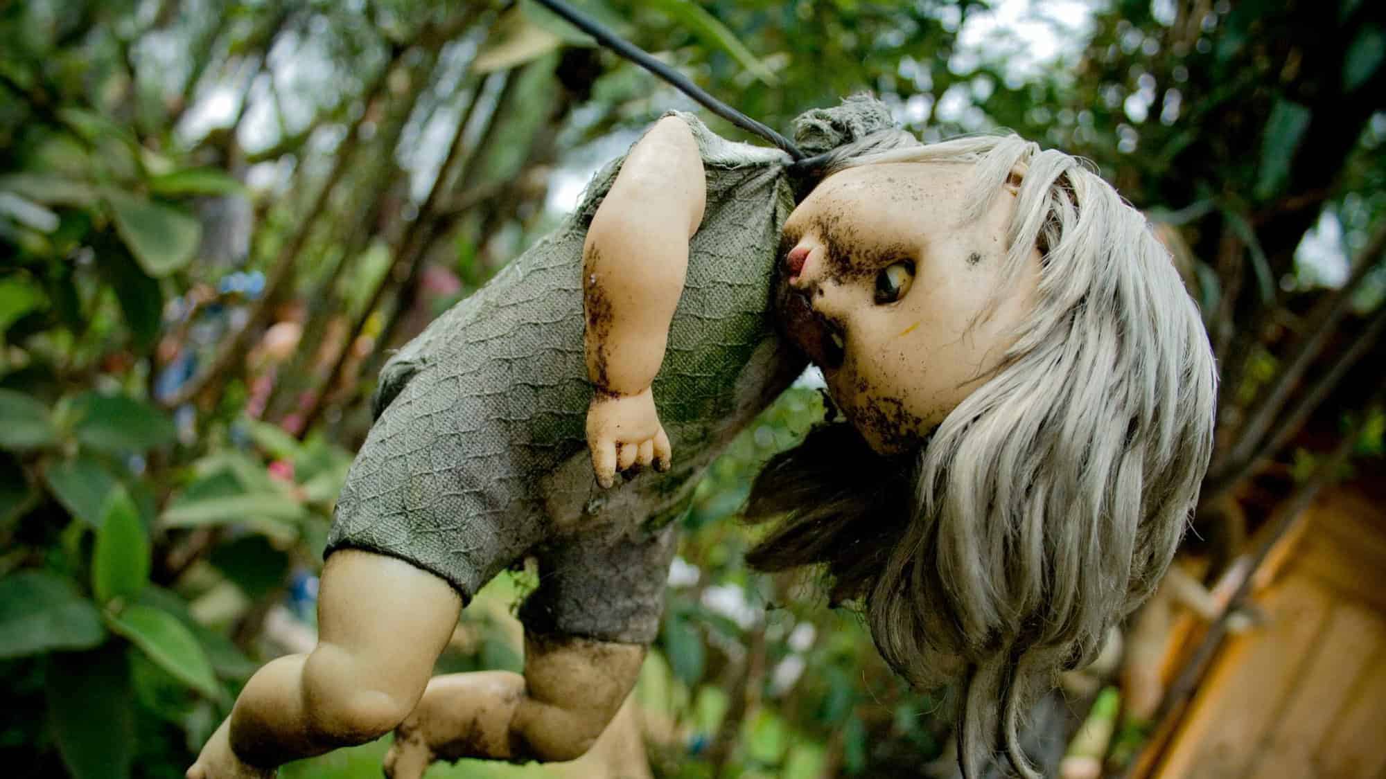 A dirty, weathered doll hanging from a string, its face cracked and body covered in grime, surrounded by trees. This unsettling image is part of the eerie collection found on the Island of the Dolls.