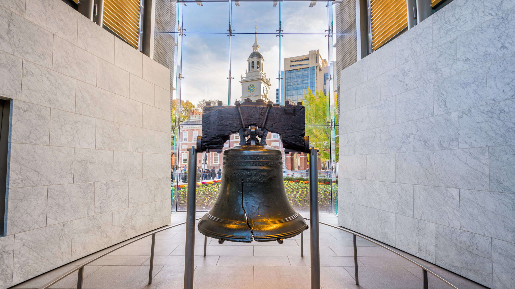 The iconic Liberty Bell displayed inside a modern structure, with the historic Independence Hall visible in the background through large glass windows.