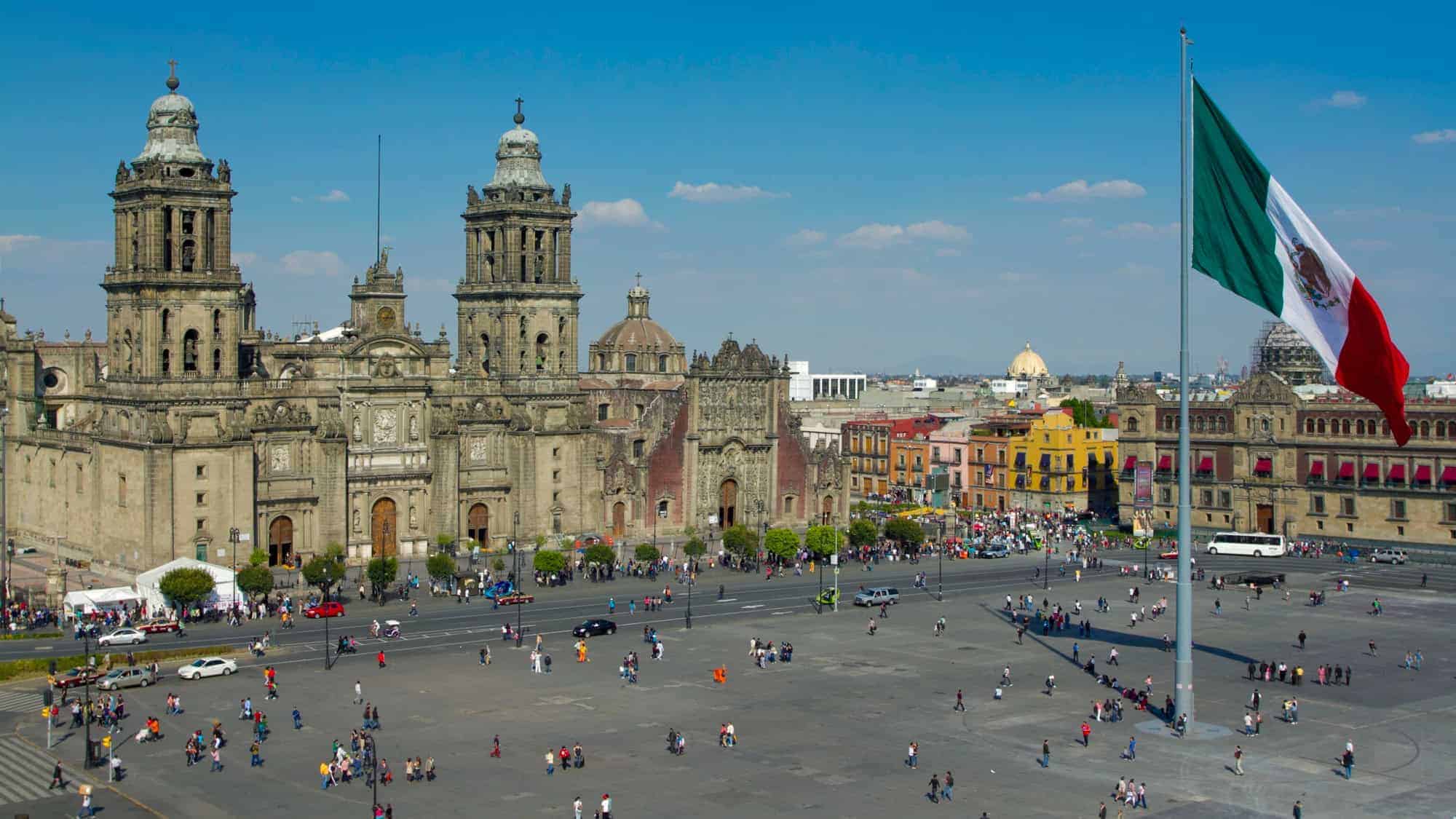 A bustling plaza in front of the Mexico City Metropolitan Cathedral, with a large Mexican flag waving prominently on the right.