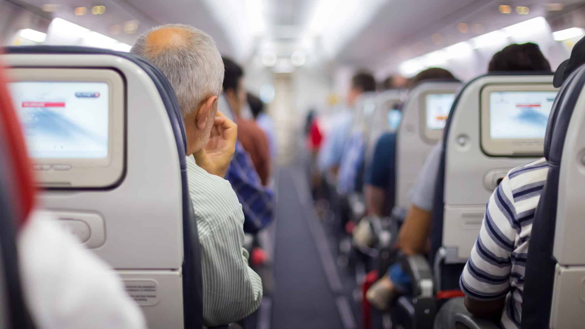 A view from the back of an airplane cabin, showing passengers seated and watching in-flight entertainment screens, with a focus on the aisle.