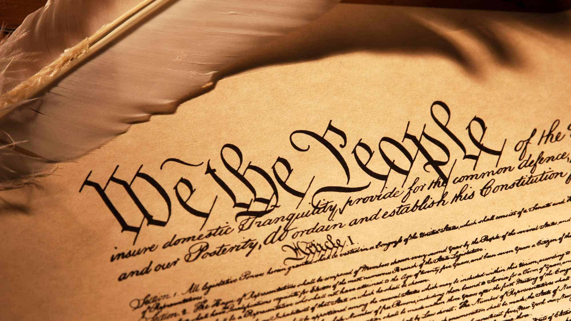 A close-up of the opening lines of the U.S. Constitution, “We the People,” written in elegant calligraphy on an old parchment, with a feather quill resting on the document.