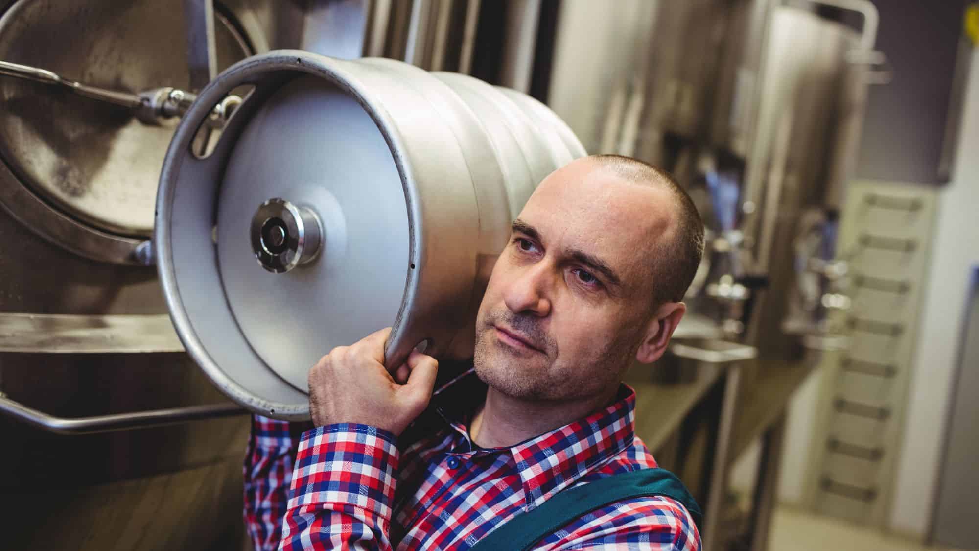A man wearing a plaid shirt carrying a metal keg on his shoulder, inside what appears to be a brewery.