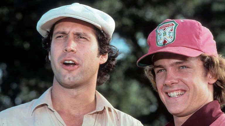 Two men, one in a white cap and the other in a red cap, are standing outside in a golf course setting, both smiling and looking off into the distance.