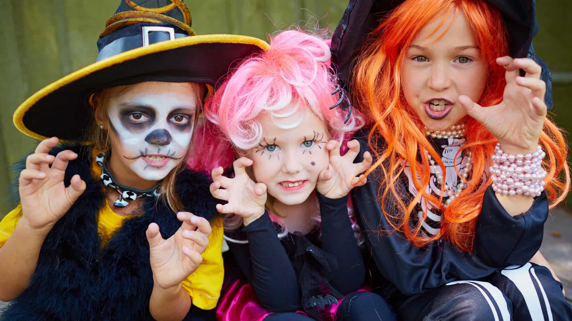 Three children dressed in colorful Halloween outfits and wigs make playful scary faces for the camera.