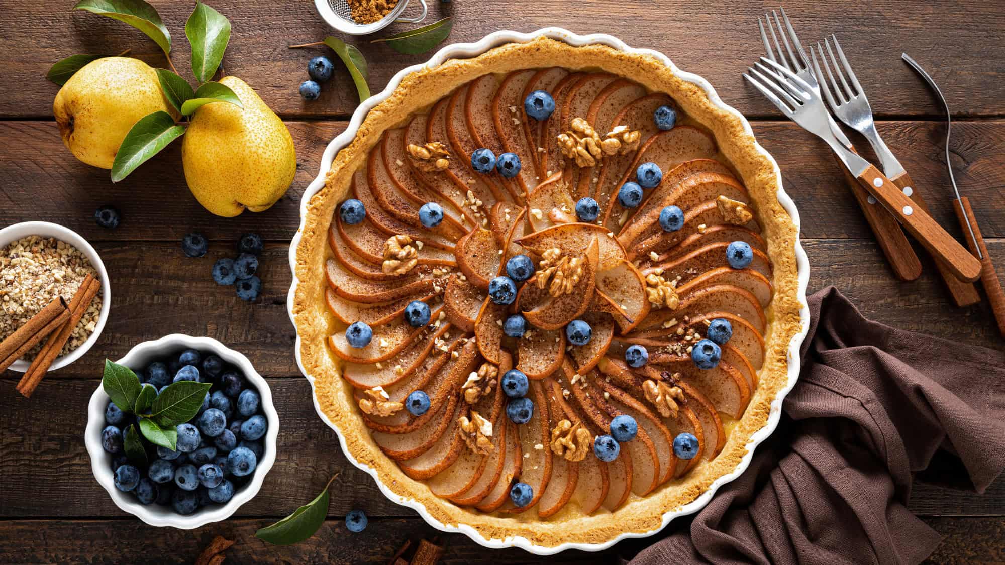A beautifully arranged tart topped with sliced pears, blueberries, and walnuts sits on a rustic wooden table.