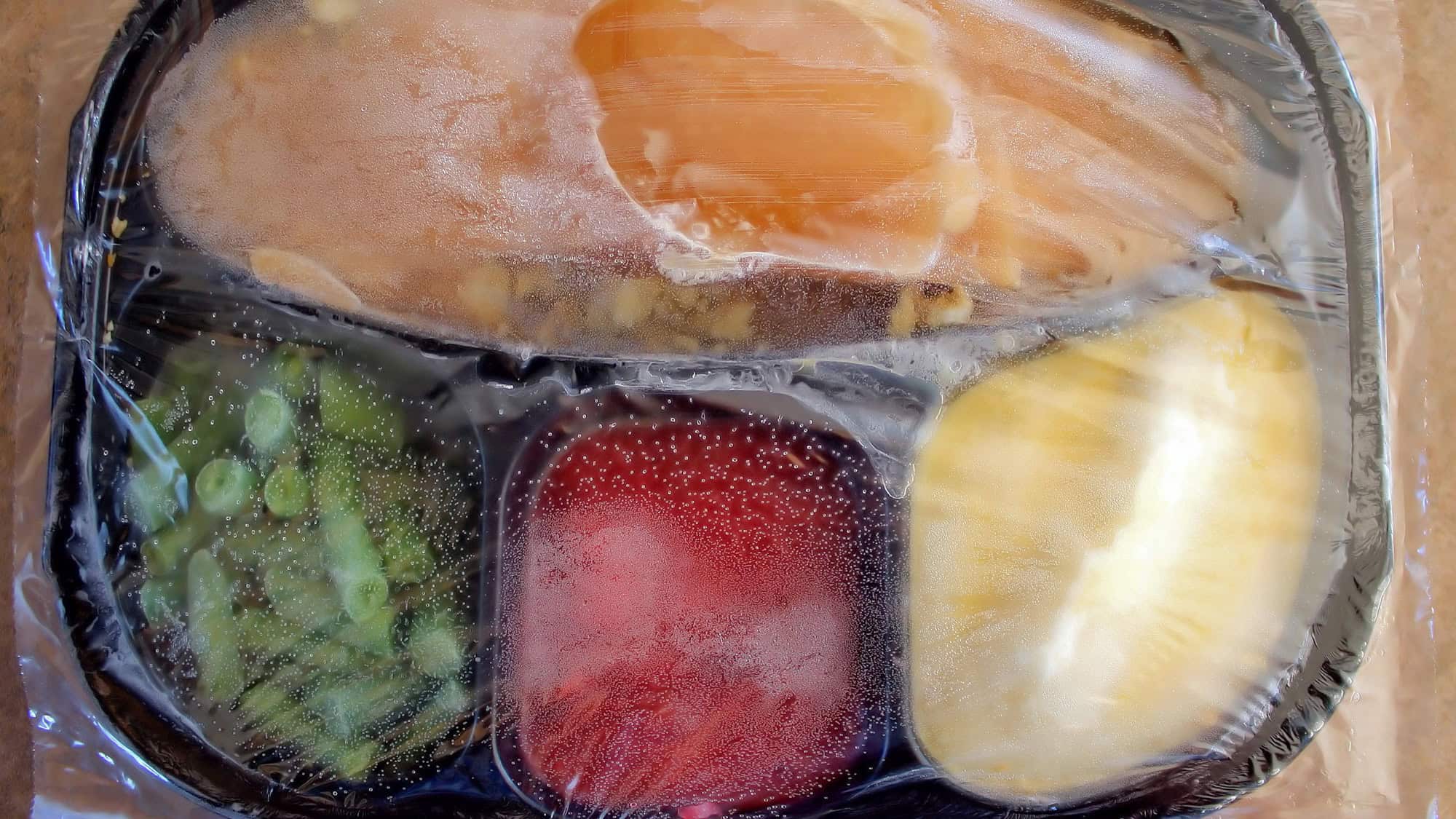 A frozen TV dinner with turkey, peas, mashed potatoes, and cranberry sauce is sealed in clear plastic.