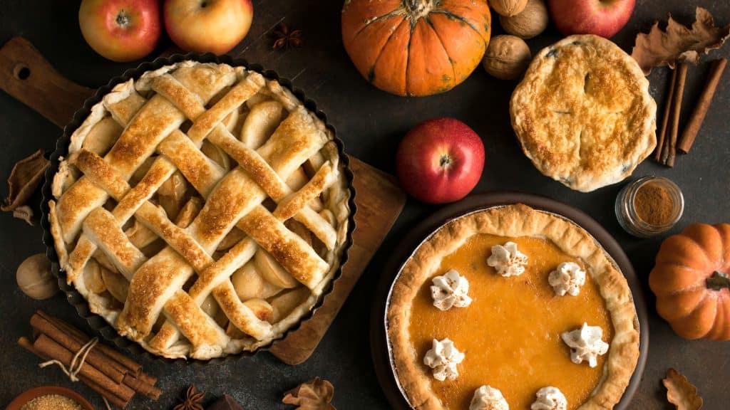 A wooden table filled with pies, including pumpikin and apple along with a few holiday decorations.