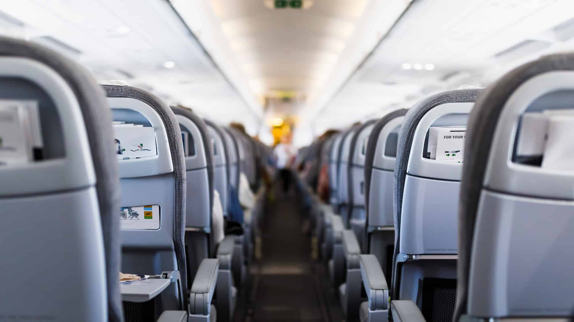 A view down the aisle of an airplane, showing rows of gray seats with tray tables and safety cards, slightly blurred toward the front.