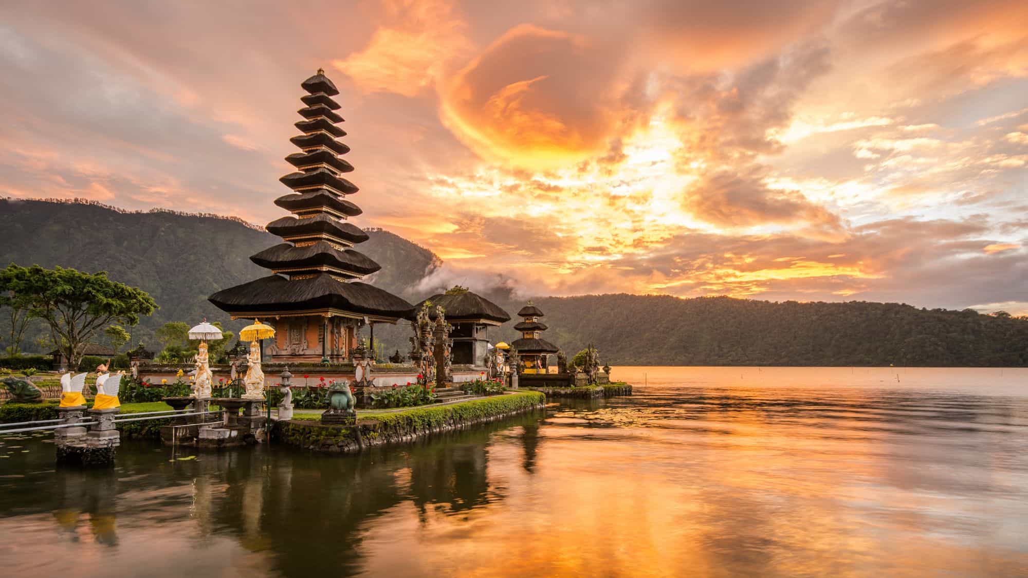 A stunning temple by a serene lake in Bali is bathed in golden hues from a dramatic sunset, with vibrant orange skies and lush green hills in the background reflecting on the water.