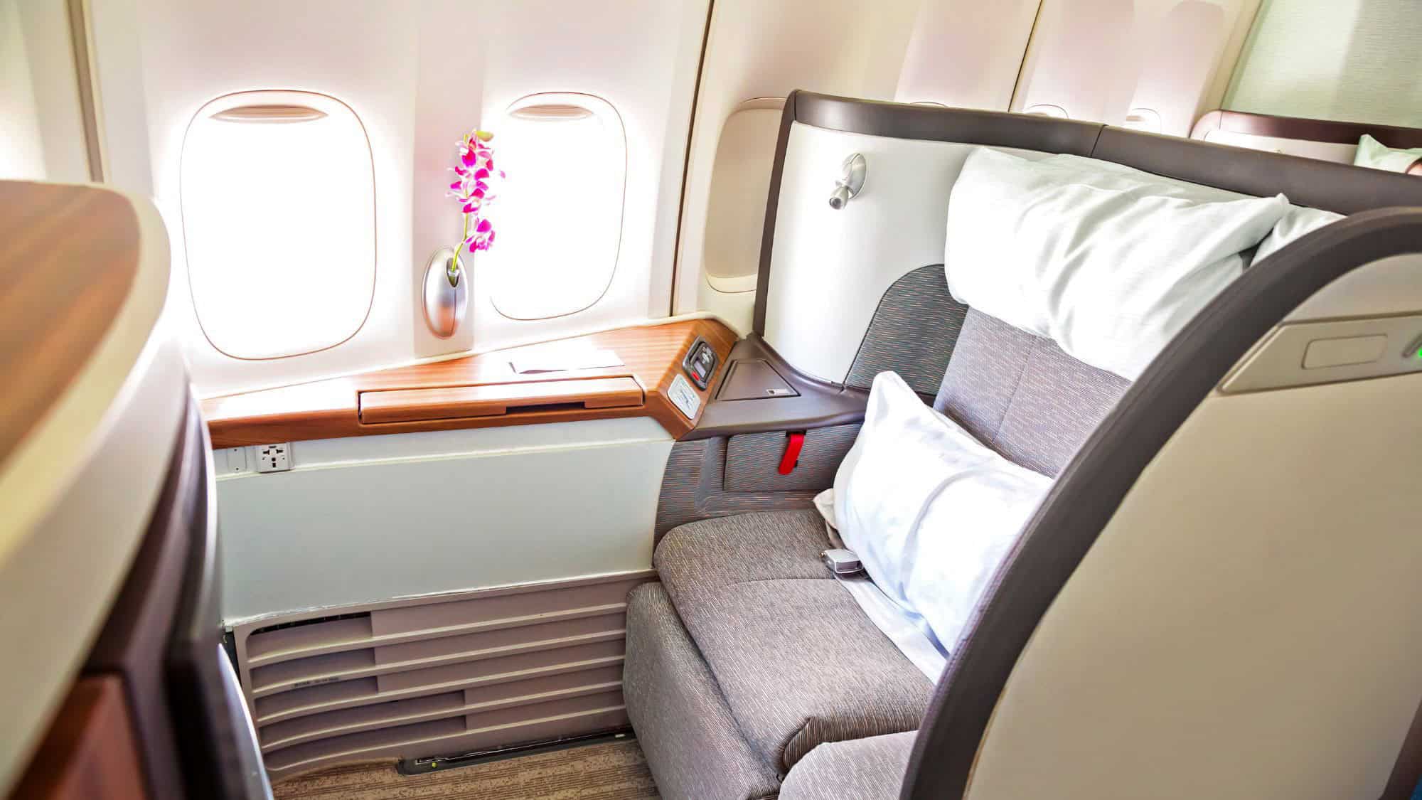 A spacious first-class airline seat with a plush pillow and wood-trimmed side table, decorated with a single orchid by the window.