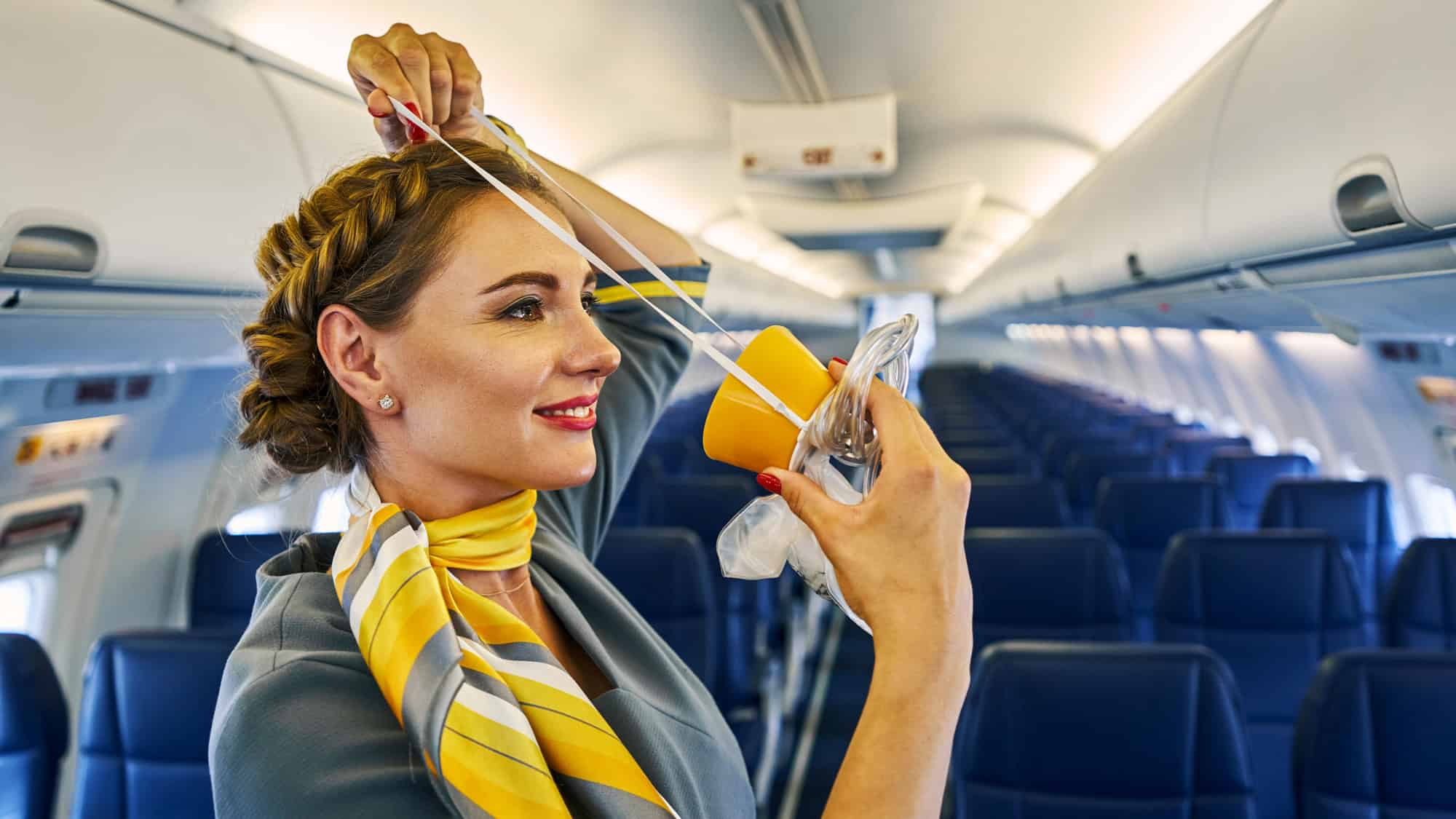 A smiling flight attendant in a yellow and gray scarf demonstrates how to place an oxygen mask over her head inside the airplane cabin.