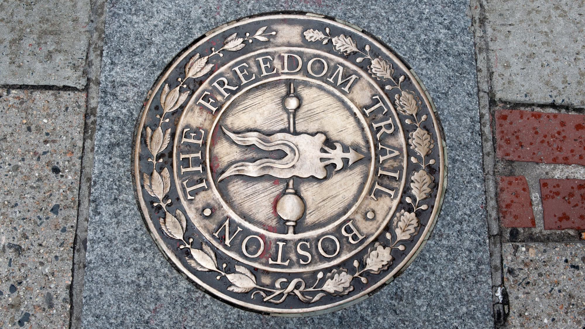 Close-up view of the bronze medallion embedded in the sidewalk marking the Freedom Trail in Boston, featuring an arrow-tipped torch and ornate laurel designs, encircled by the words "The Freedom Trail Boston."