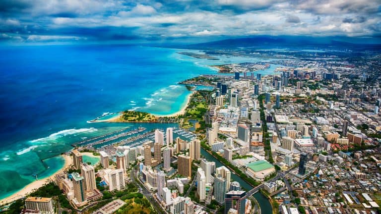 An aerial view of Honolulu, Hawaii, featuring turquoise ocean waters, white sandy beaches, and a dense skyline of high-rise buildings along the coastline.
