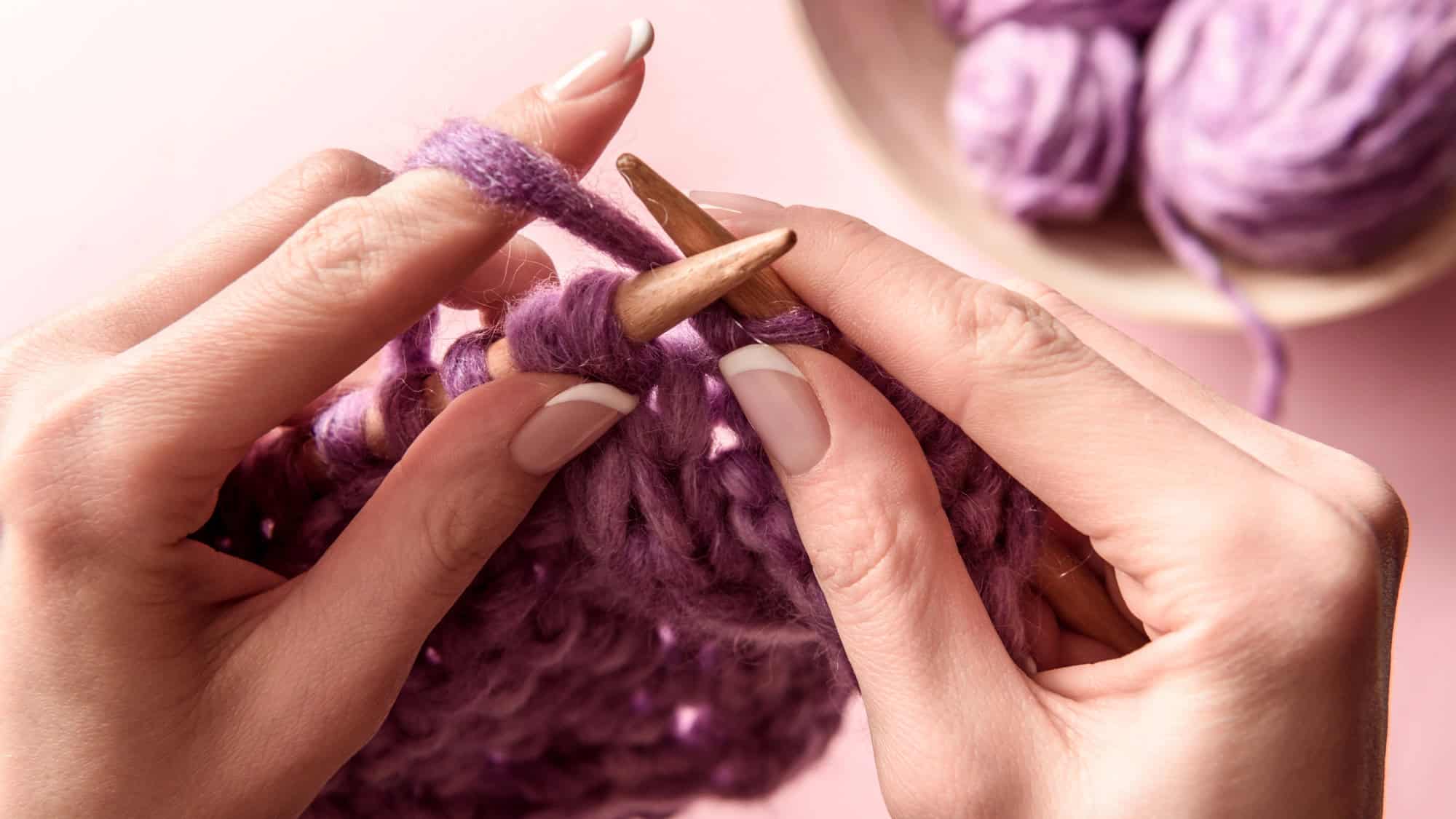 Close-up of hands using wooden knitting needles to work with thick purple yarn, with a bowl of yarn in the background.