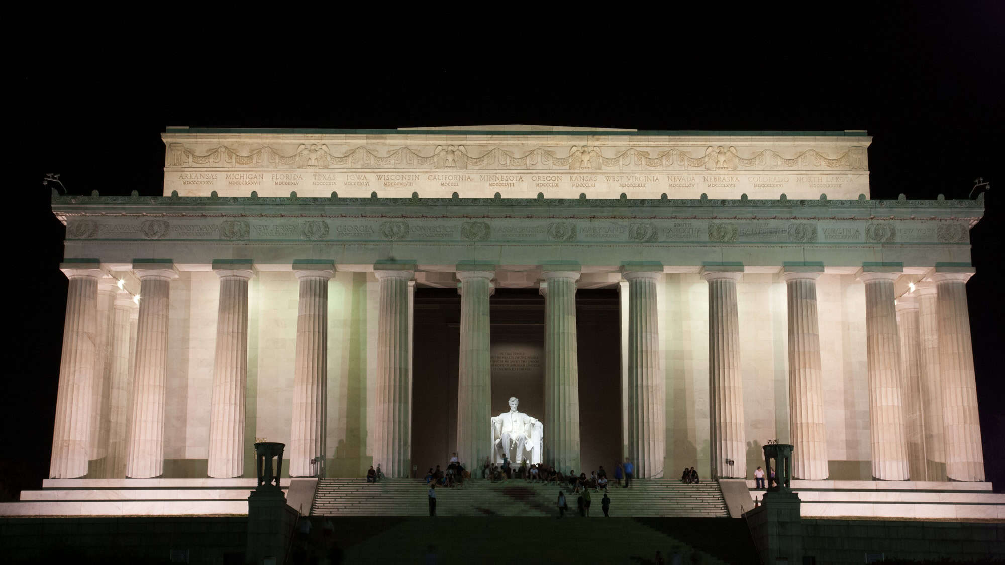 The Lincoln Memorial in Washington, D.C., lit up against a dark night sky, with its grand neoclassical columns glowing and the statue of Abraham Lincoln illuminated at the center, surrounded by evening visitors.