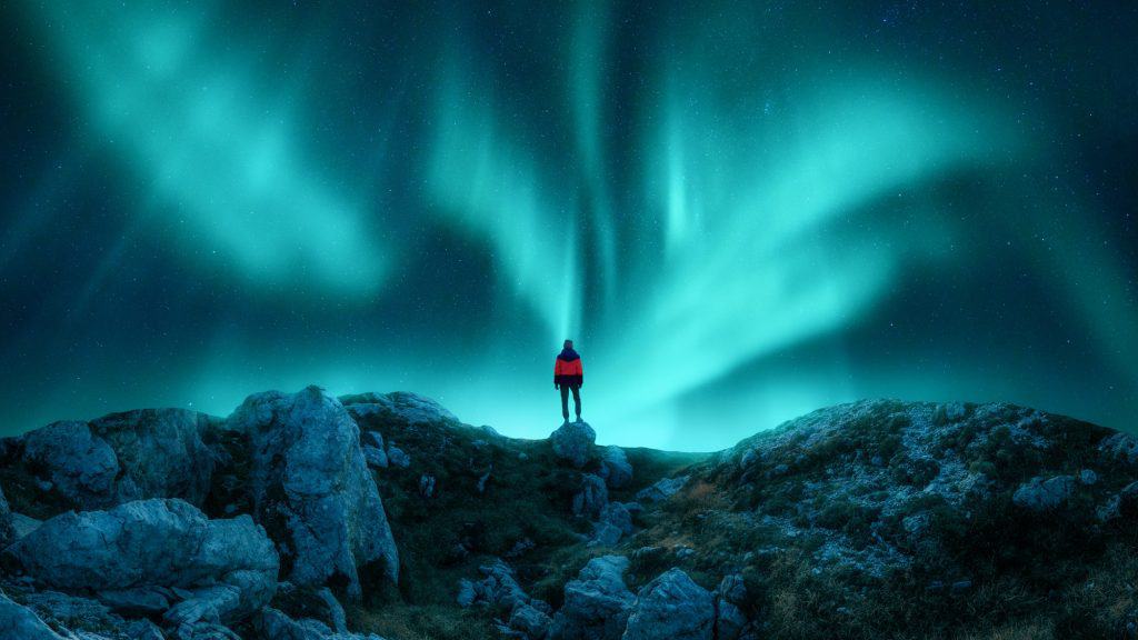 A person in a red jacket stands atop rocky terrain, silhouetted against a dramatic green aurora illuminating the night.