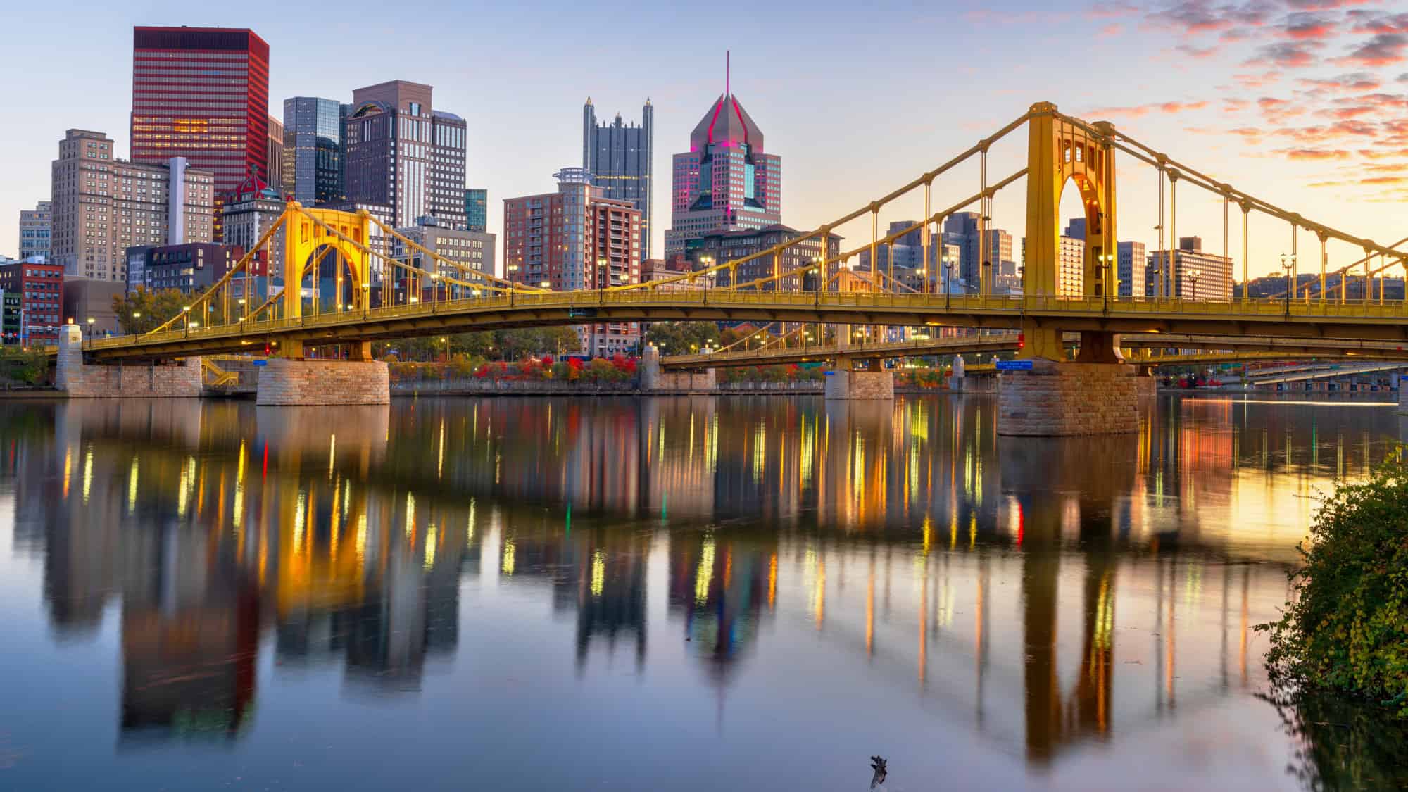 Golden hour lights up the Roberto Clemente Bridge and downtown Pittsburgh, casting colorful reflections across the still waters of the Allegheny River.