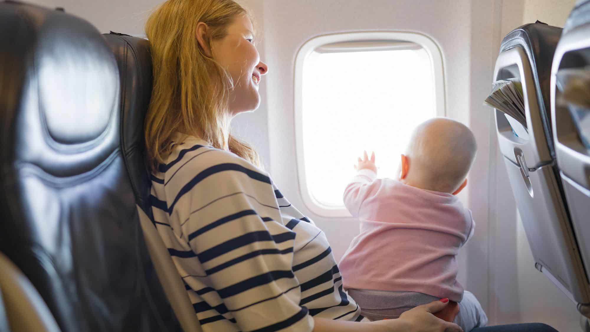 A smiling woman holds a baby on her lap in an airplane seat as the child reaches toward the bright window.