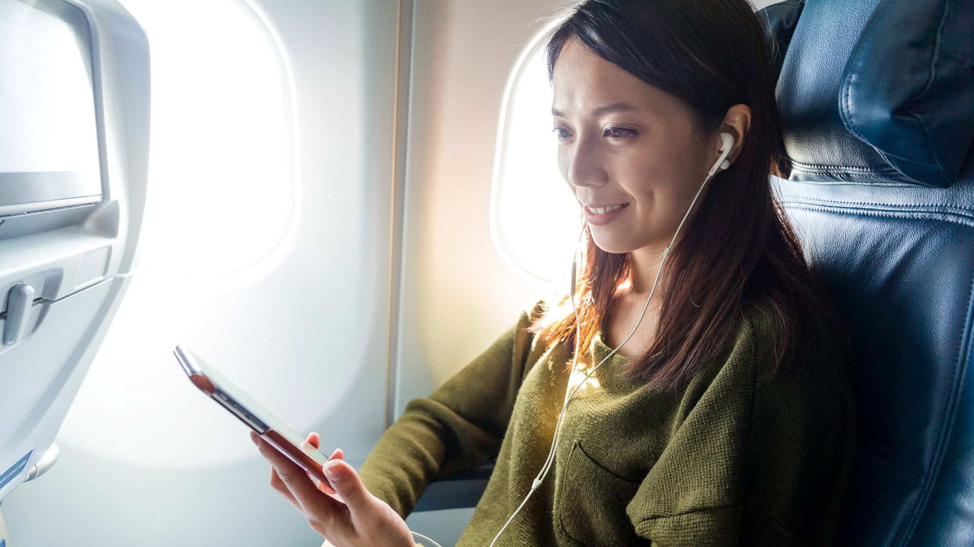 A woman in a green sweater sits by the airplane window, smiling while looking at her phone and listening through wired earbuds.