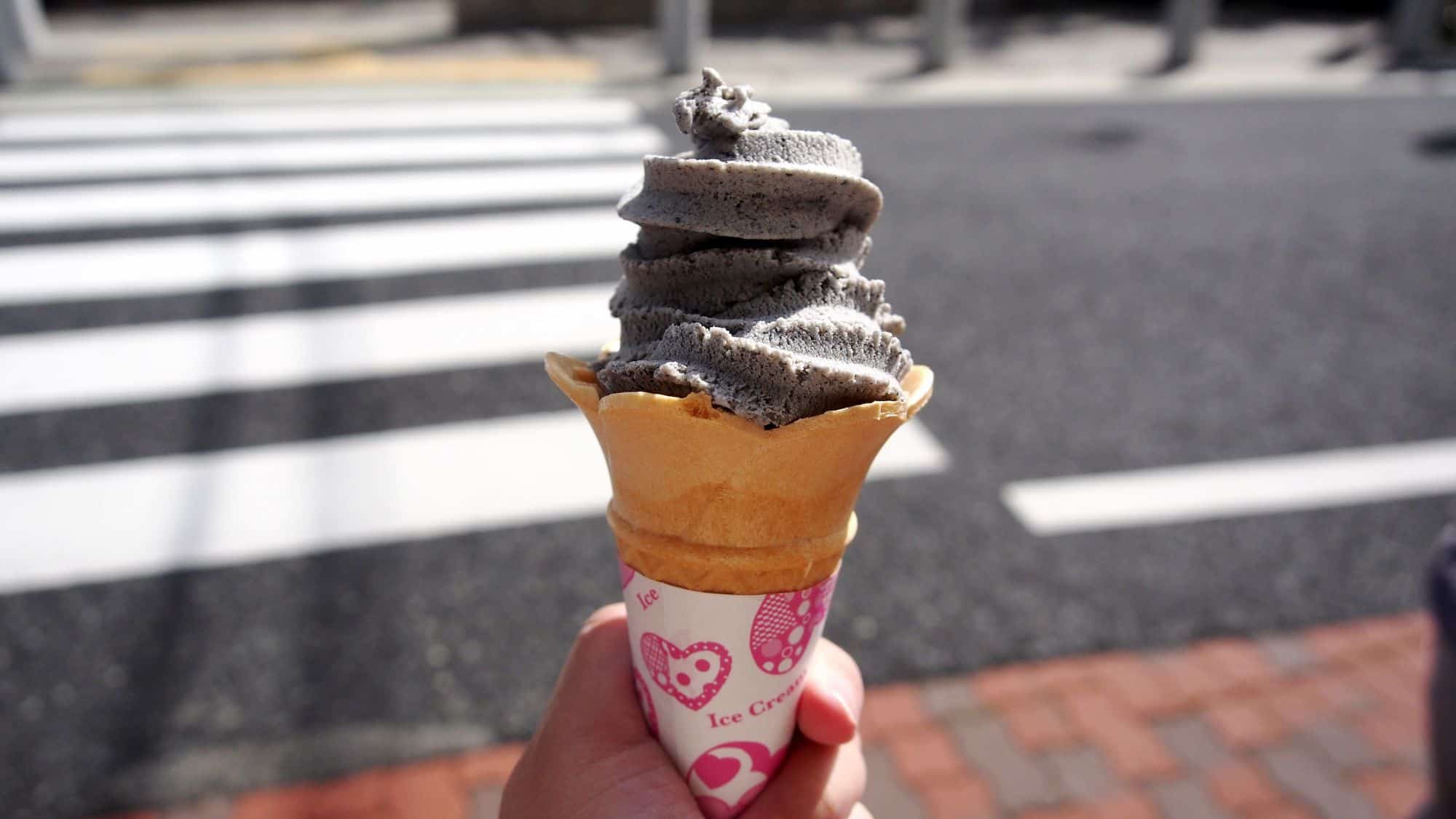 A single cone of black sesame soft-serve ice cream with a deep charcoal hue, held against a blurred city street background with pedestrian crosswalk lines.
