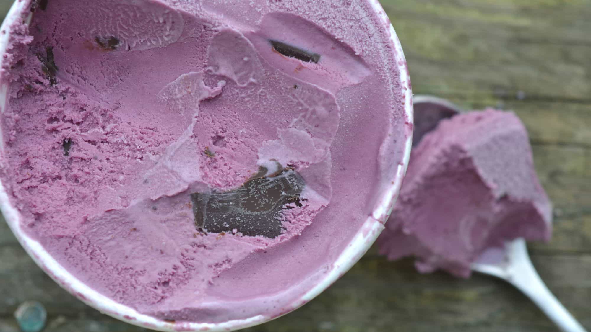 A top-down view of a container filled with rich, purple-black raspberry ice cream with visible chocolate chunks, accompanied by a scoop resting on a rustic wooden surface.