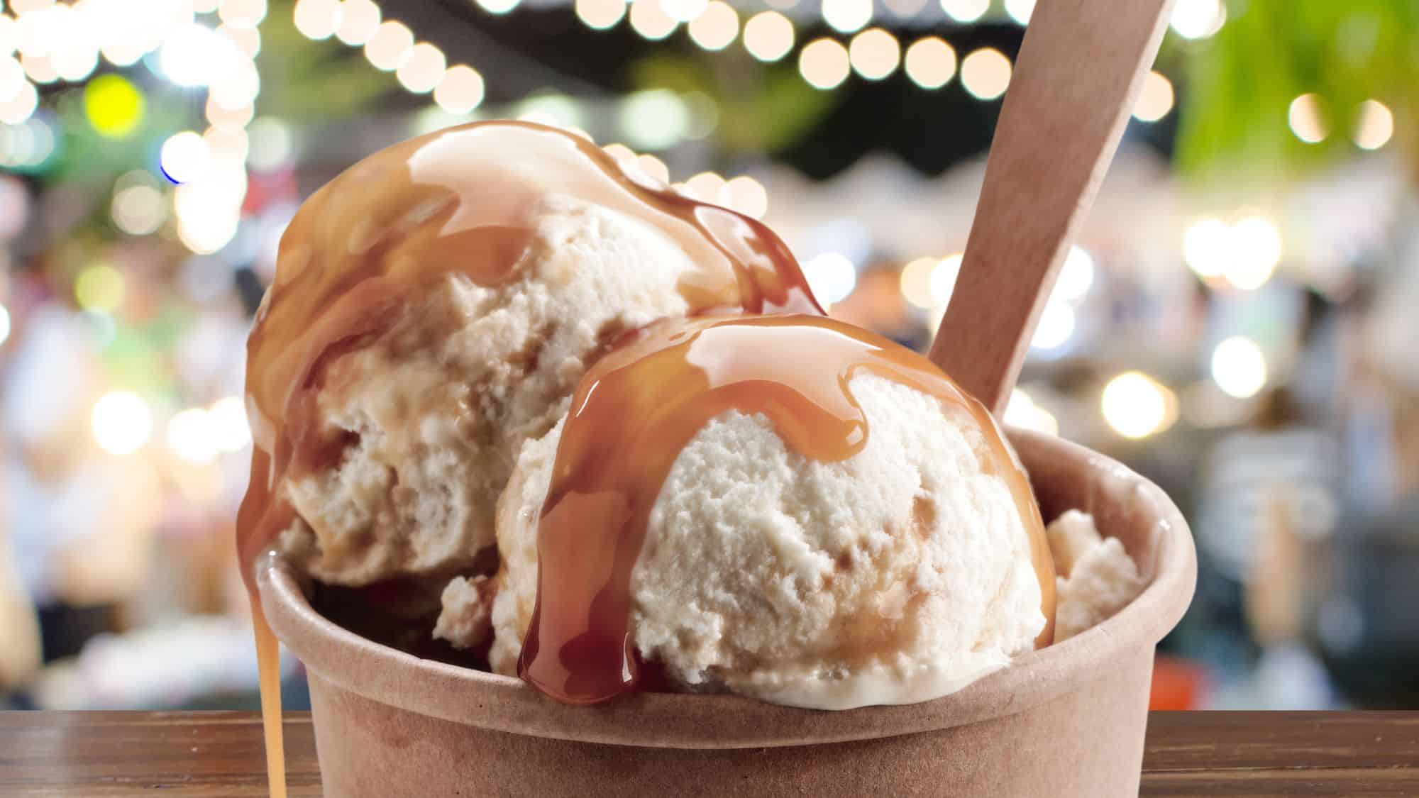 A cup of vanilla ice cream drizzled with caramel sauce, served in a paper cup with a wooden spoon. The background features blurred lights, adding to the festive atmosphere.