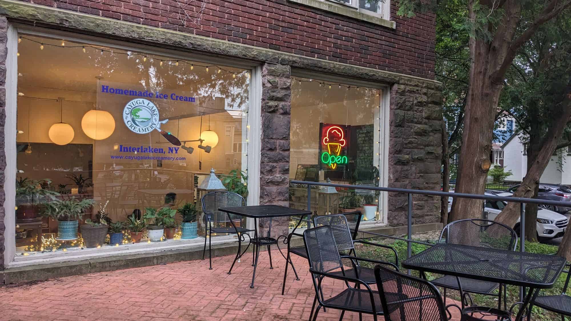 A cozy ice cream shop with large windows showing the warmly lit interior, potted plants, and a neon "Open" sign featuring an ice cream cone. The exterior has a brick and stone facade with outdoor seating.