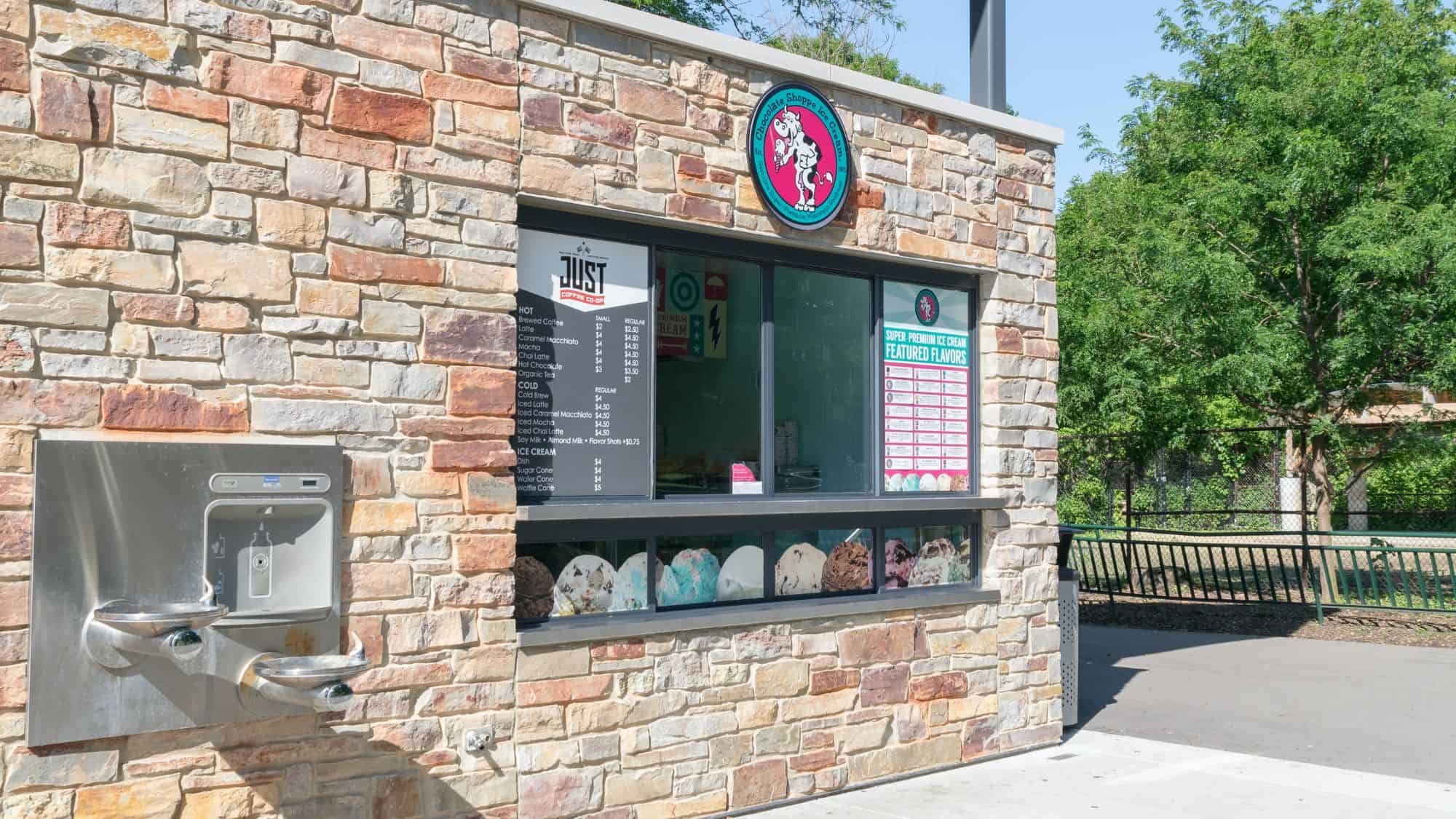 A small ice cream stand with a rustic stone facade featuring an array of displayed ice cream flavors and a menu board. A water fountain is mounted on the wall next to the shop.