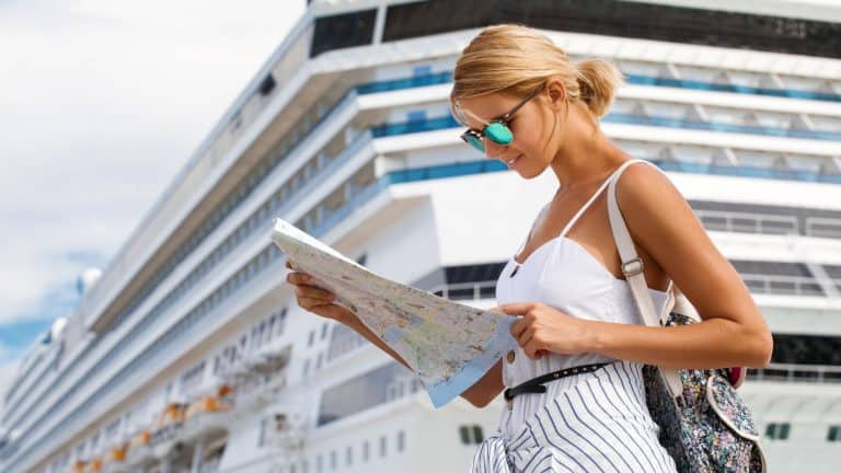 A woman in a stylish summer outfit and sunglasses studies a map near a large cruise ship docked in the background, preparing for an adventure at her next port of call.