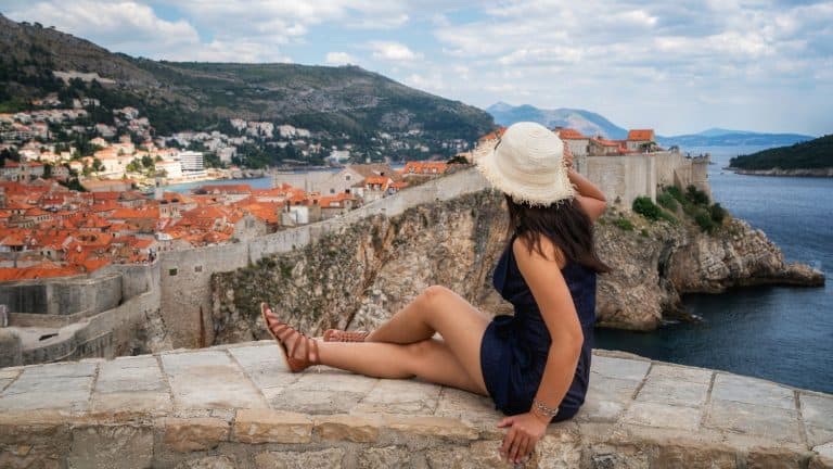 A woman in a sun hat sits on a stone ledge overlooking the iconic terracotta rooftops and fortress walls of Dubrovnik, Croatia, with the Adriatic Sea and distant hills in the background.
