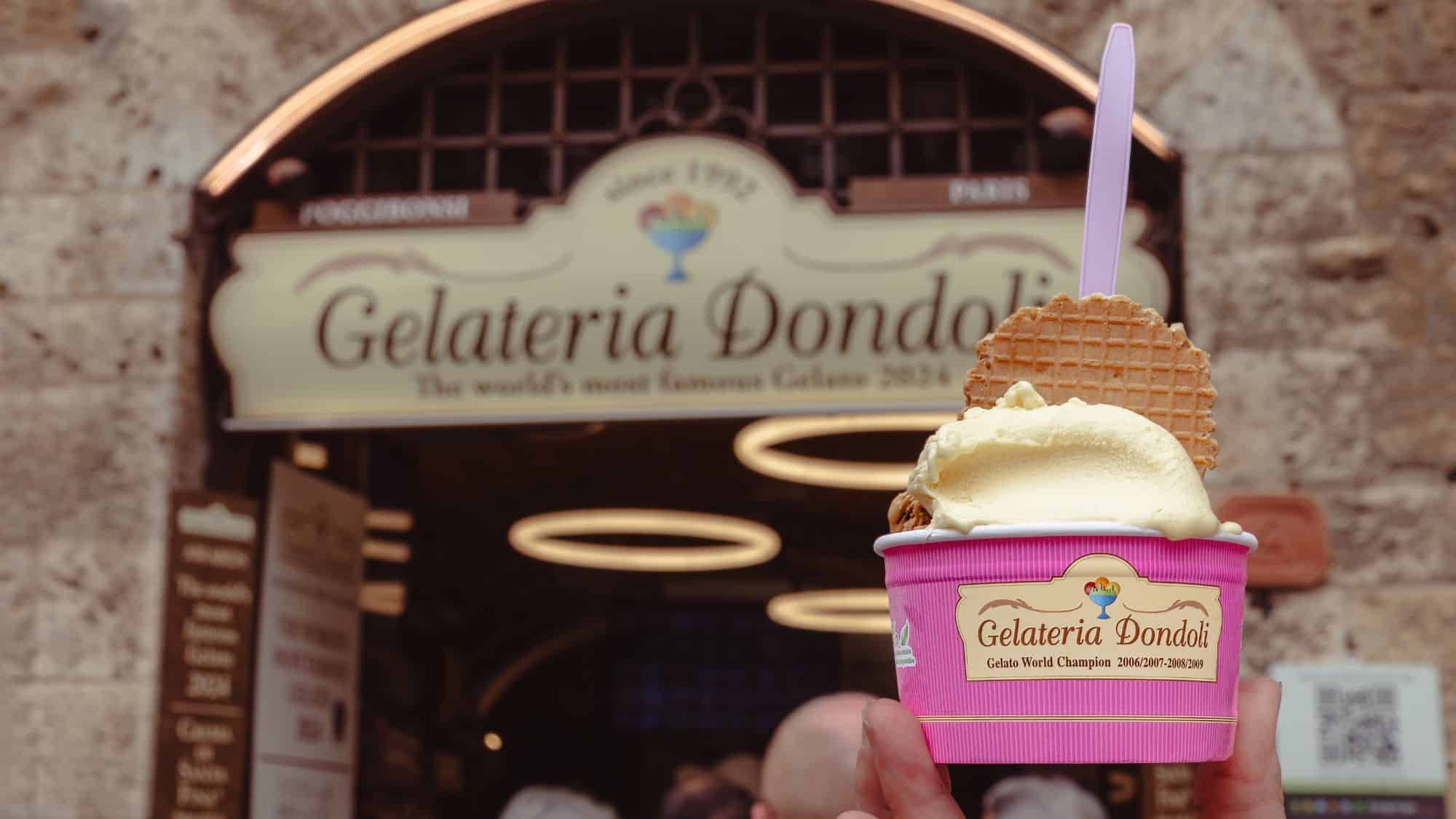 A hand holding a cup of gelato with a waffle cookie in front of the famous Gelateria Dondoli shop sign. The store's signage highlights its recognition as a world champion gelato shop.