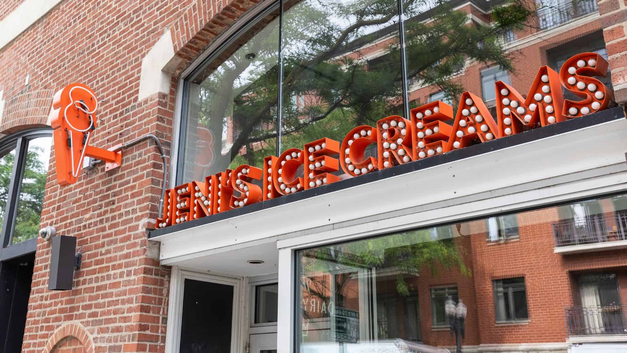 The exterior of Jeni’s Ice Cream shop with a bright orange retro-style neon sign on a brick building. The large windows reflect the urban surroundings.