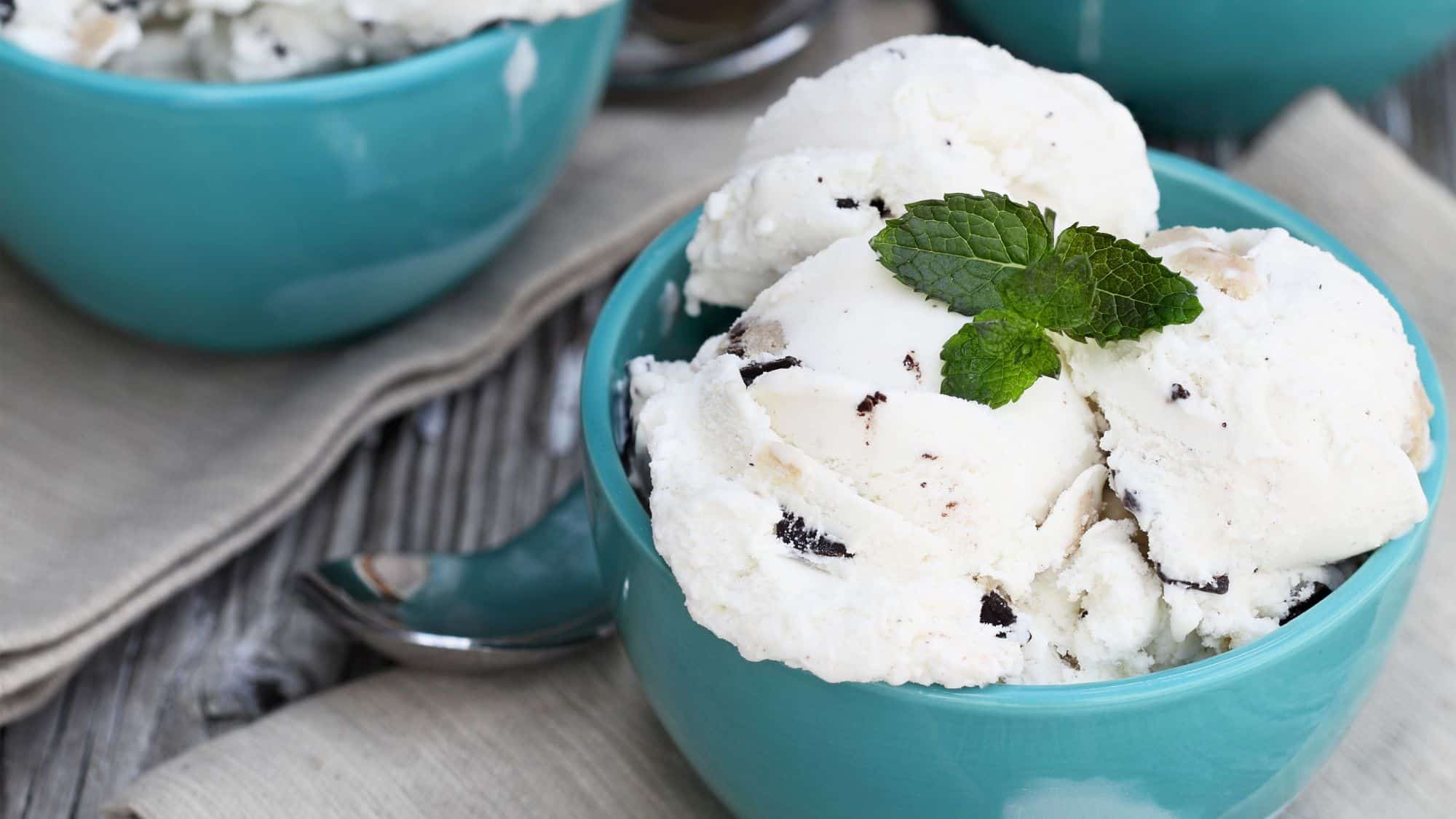 A close-up of a bowl of creamy white ice cream with dark chocolate chips, garnished with a fresh mint leaf. The ice cream is served in a stylish turquoise bowl placed on a rustic wooden table.