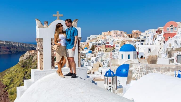 A couple stands on a whitewashed rooftop with the blue-domed churches and Aegean Sea in the background. The sunny sky and iconic Cycladic architecture create a romantic and breathtaking scene.