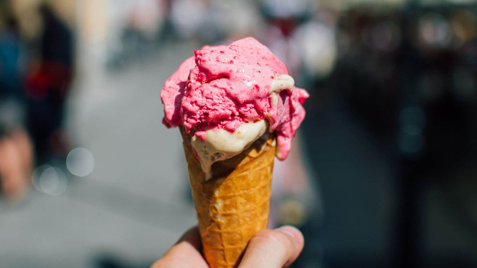 A hand holding a waffle cone topped with a scoop of pink strawberry ice cream, melting slightly. The blurred background shows a busy outdoor setting.